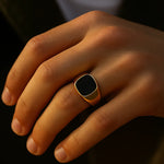 Herren-Siegelring aus vergoldetem Edelstahl mit schwarzem Achat, getragen am Ringfinger; Nahaufnahme im warmen Fensterlicht, natürliche Hauttöne, moderner Look.
