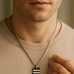 Nahaufnahme eines Mannes mit Herren-Halskette aus Edelstahl; schwarzer Carbon-Anhänger mit zwei silbernen Streifen, getragen über beigem Pullover, weiches Fensterlicht.
