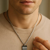 Nahaufnahme eines Mannes mit Herren-Halskette aus Edelstahl; schwarzer Carbon-Anhänger mit zwei silbernen Streifen, getragen über beigem Pullover, weiches Fensterlicht.
