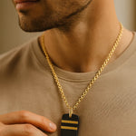 Herren-Halskette: vergoldete Edelstahlkette mit schwarzem Dogtag und zwei goldenen Streifen, getragen am Hals im warmen Fensterlicht.
