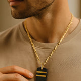 Herren-Halskette: vergoldete Edelstahlkette mit schwarzem Dogtag und zwei goldenen Streifen, getragen am Hals im warmen Fensterlicht.
