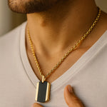 Herren-Halskette: vergoldete Edelstahlkette mit zweifarbigem Dogtag (schwarz/gold), getragen am Hals eines Mannes im warmen Fensterlicht, nah aufgenommen.
