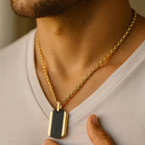 Herren-Halskette: vergoldete Edelstahlkette mit zweifarbigem Dogtag (schwarz/gold), getragen am Hals eines Mannes im warmen Fensterlicht, nah aufgenommen.
