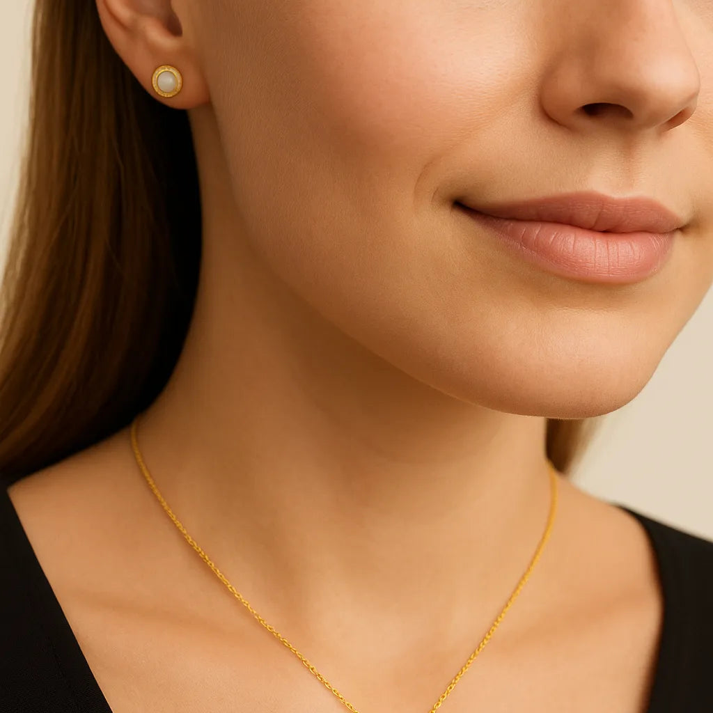 Nahaufnahme einer Frau mit cremefarbenem Hintergrund, die eine zarte goldene Halskette mit Cabochon-Perlenanhänger und dezentem Zirkonia-Kranz trägt; ein passender Ohrring ist auf einer Seite sichtbar. Der Schmuck wirkt matt-edel und elegant.
