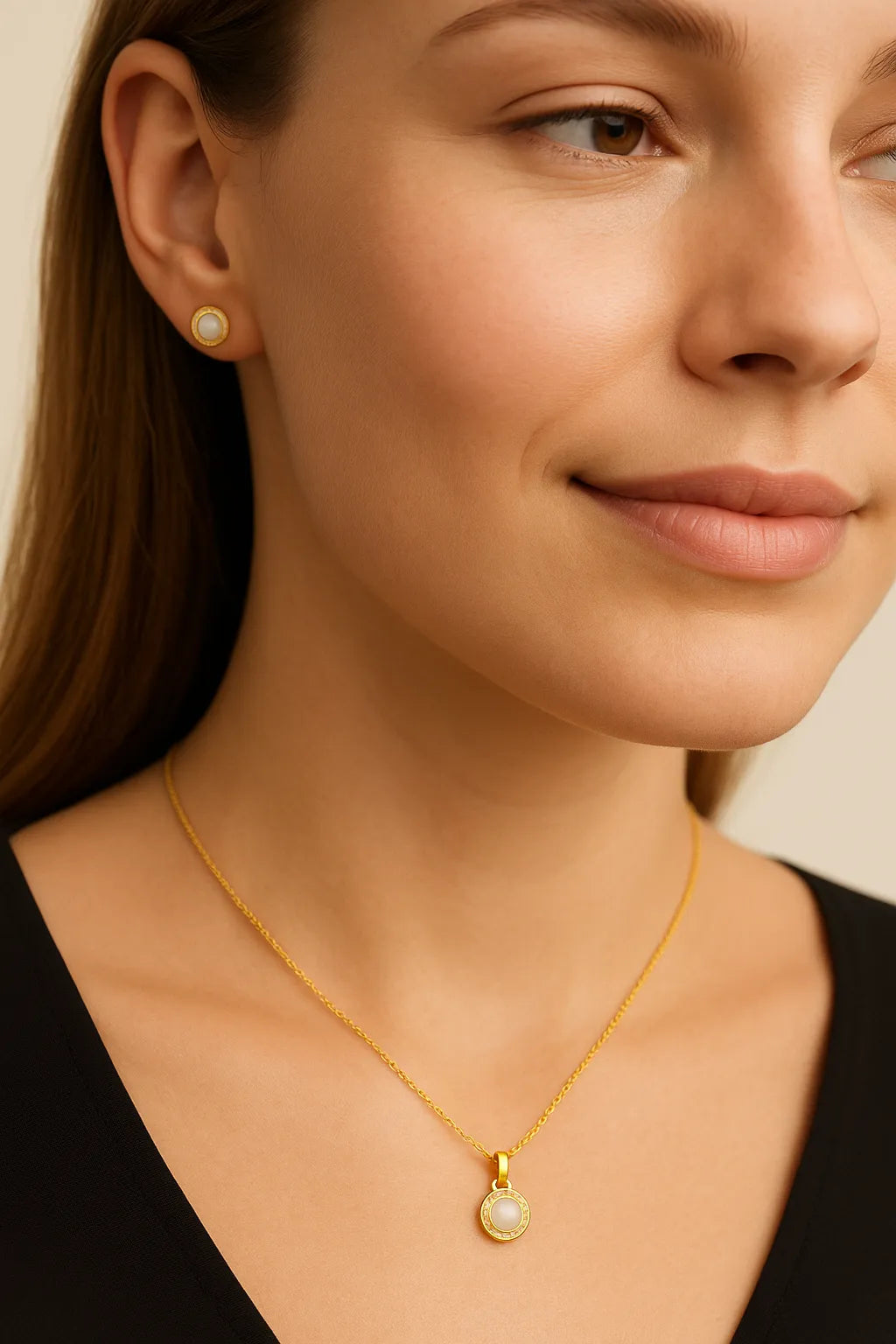 Nahaufnahme einer Frau mit cremefarbenem Hintergrund, die eine zarte goldene Halskette mit Cabochon-Perlenanhänger und dezentem Zirkonia-Kranz trägt; ein passender Ohrring ist auf einer Seite sichtbar. Der Schmuck wirkt matt-edel und elegant.
