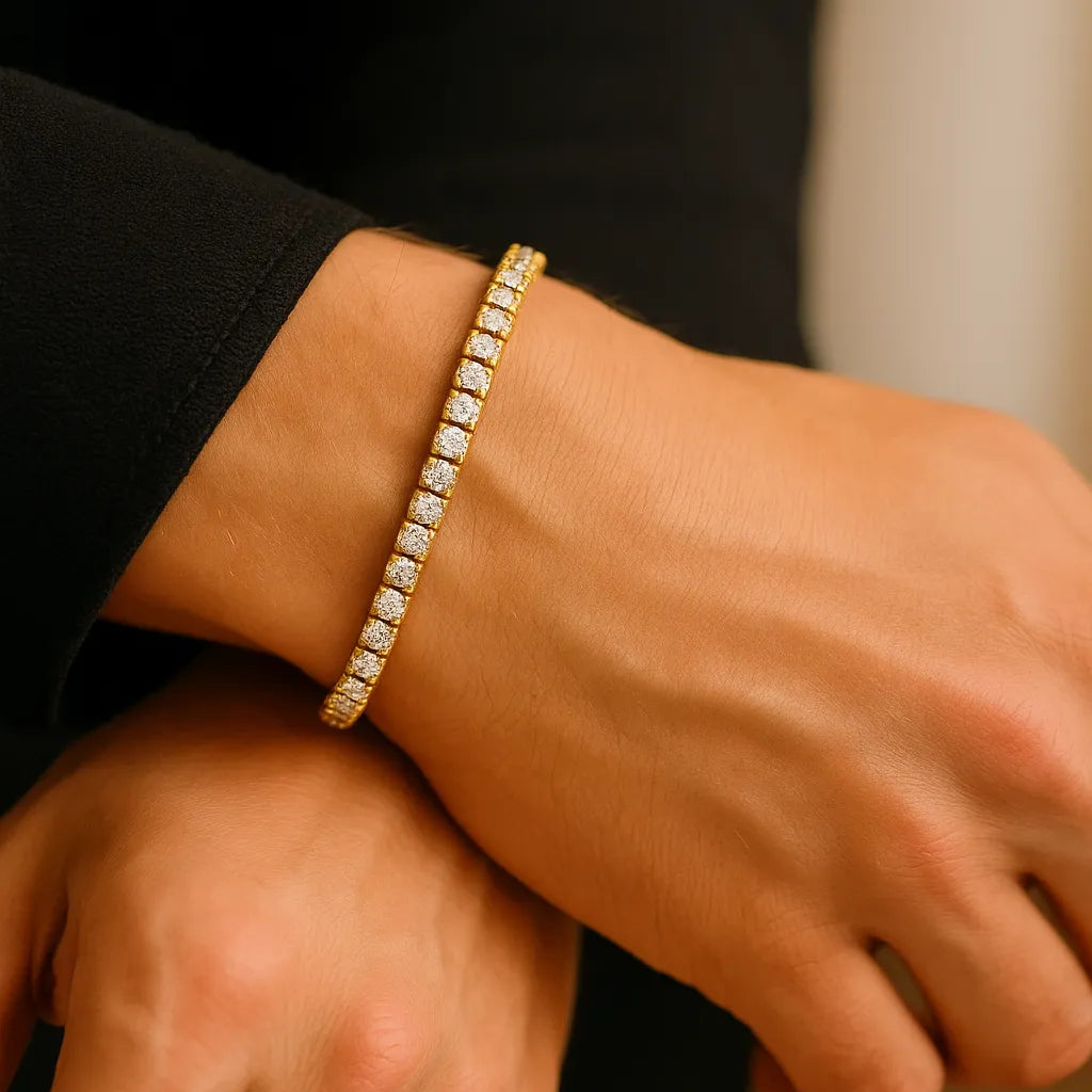 Nahaufnahme eines Männerhandgelenks mit goldenem Tennisarmband aus Edelstahl (18K-IPG) und funkelnden Zirkonia, getragen zu schwarzem Ärmel, weiches Golden-Hour-Fensterlicht.
