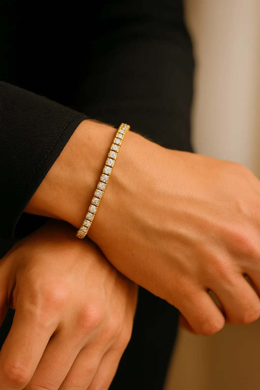 Nahaufnahme eines Männerhandgelenks mit goldenem Tennisarmband aus Edelstahl (18K-IPG) und funkelnden Zirkonia, getragen zu schwarzem Ärmel, weiches Golden-Hour-Fensterlicht.
