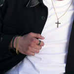 Herren-Armband aus vergoldetem Edelstahl (IPG) am Handgelenk, kombiniert mit Lederarmband, Bicolor-Ring und goldener Kreuzkette; schwarzer Mantel, weißes T-Shirt.
