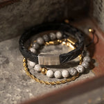 **„Son of Noa Herren-Armbänder im Layering-Look – schwarzes Lederarmband, graues Perlenarmband und goldene Kette auf rustikalem Untergrund.“**
