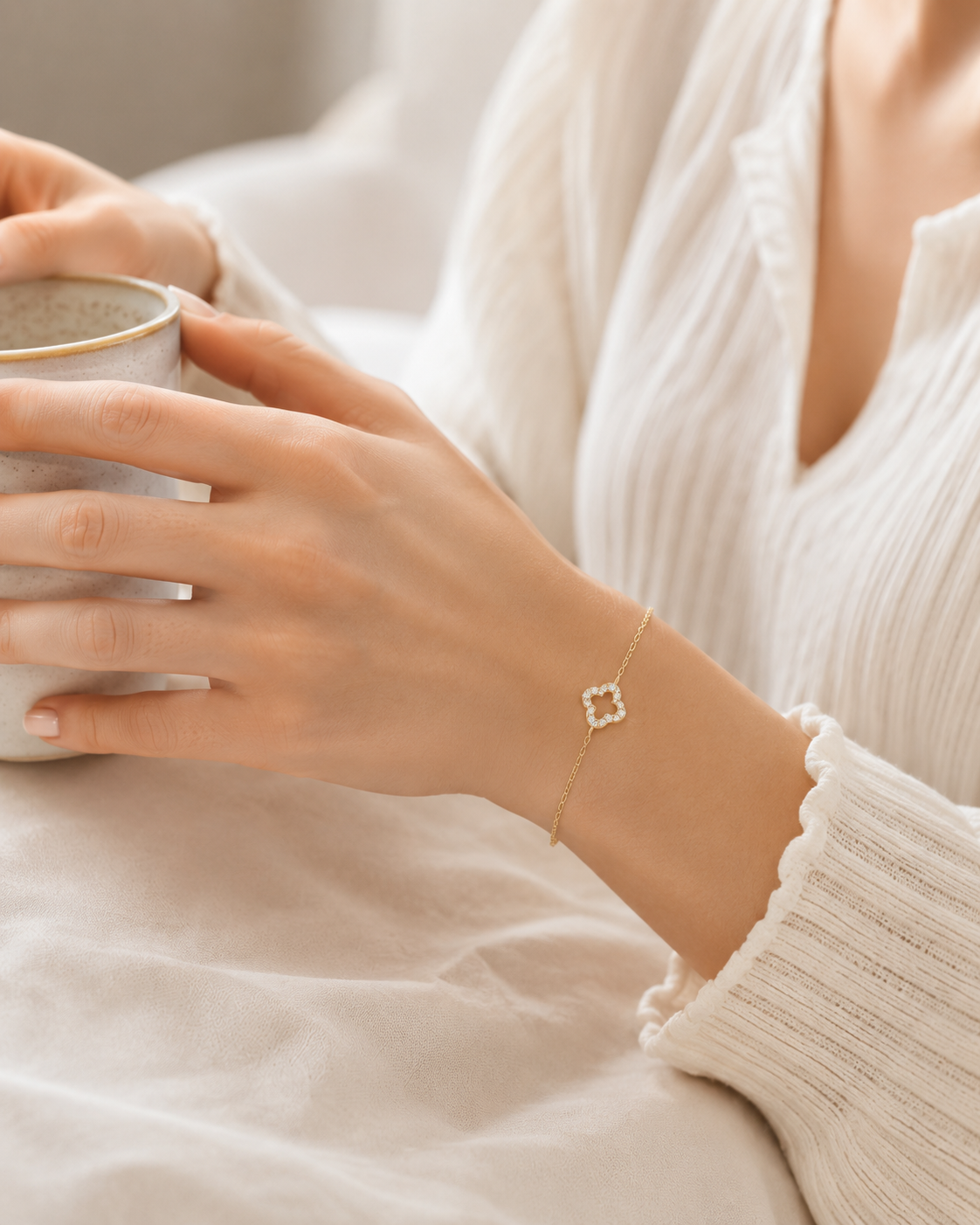 Vergoldetes PamelaNor Armband mit feinem Kleeblatt-Detail, getragen zu heller Bluse beim Halten einer Keramiktasse.