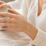 Vergoldetes PamelaNor Armband mit feinem Kleeblatt-Detail, getragen zu heller Bluse beim Halten einer Keramiktasse.