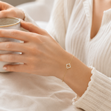 Vergoldetes PamelaNor Armband mit feinem Kleeblatt-Detail, getragen zu heller Bluse beim Halten einer Keramiktasse.