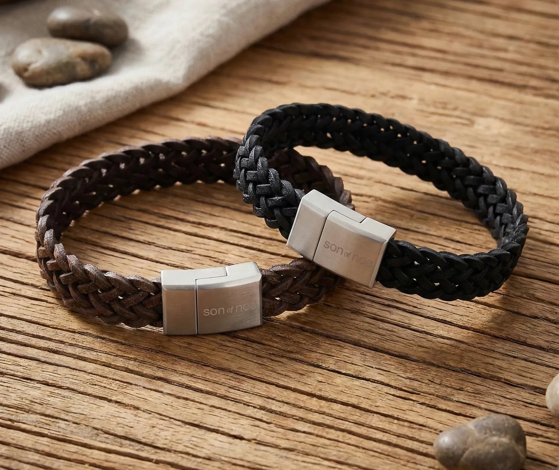 „Zwei Herren-Lederarmbänder von Son of Noa im schwarzen und braunen Fischgrätenmuster mit Edelstahl-Magnetverschluss auf rustikalem Holztisch.“
