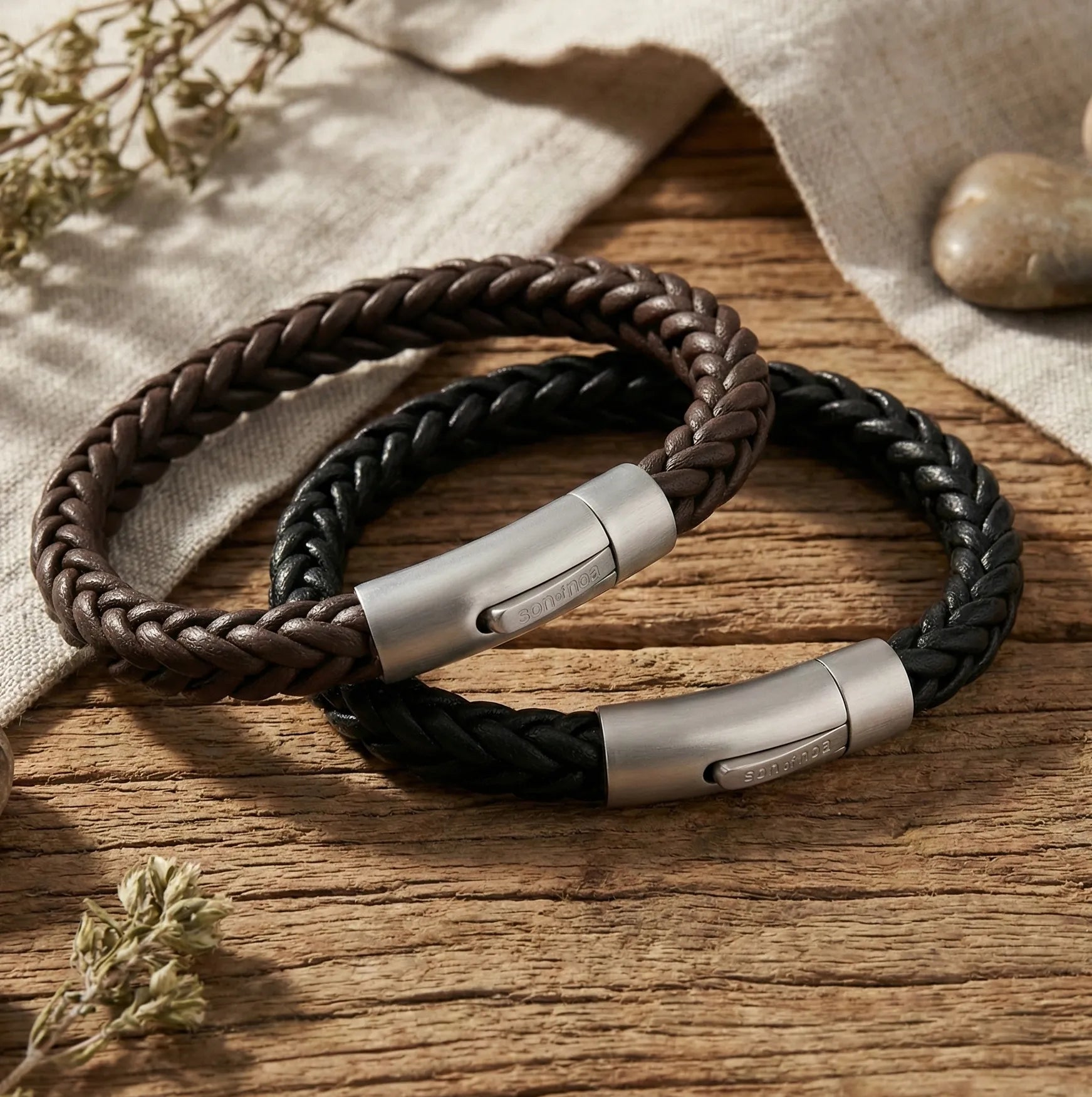 „Zwei rundgeflochtene Herren-Lederarmbänder in Braun und Schwarz mit mattem Edelstahlverschluss von Son of Noa auf rustikalem Holztisch.“
