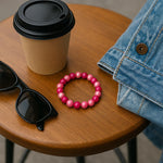 Ein stimmungsvolles Stillleben zeigt ein pink-rosa Perlenarmband auf einem Holztisch in urbaner Umgebung. Daneben liegen eine schwarze Sonnenbrille, ein Coffee-to-go-Becher und eine Jeansjacke – alles in natürlichem Tageslicht arrangiert.