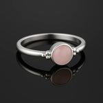 Eleganter Silberring mit zart rosafarbenem Rosenquarz Cabochon und seitlichen Silberkügelchen, auf einem dunklen, spiegelnden Hintergrund platziert; der Ring spiegelt sich klar unterhalb.