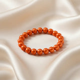 Ein Armbands aus glänzenden, orangefarbenen Perlen, das auf einem beigefarbenen, seidenartigen Stoff liegt.