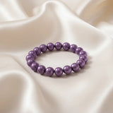 Ein Armbands aus glänzenden, lila Perlen, das auf einem beigefarbenen, seidenartigen Stoff liegt.