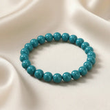 Ein Armbands aus glänzenden, türkisfarbenen Perlen, das auf einem beigefarbenen, seidenartigen Stoff liegt.