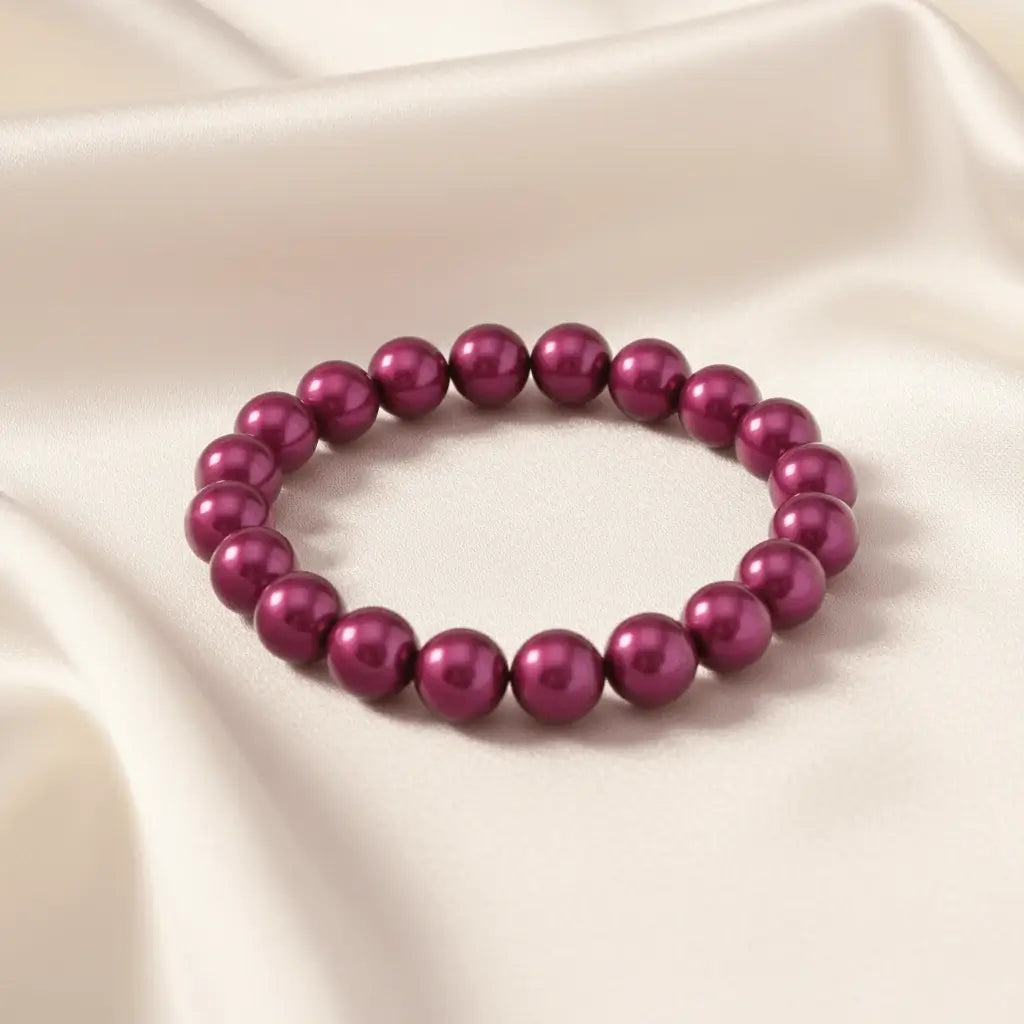 Ein Armbands aus glänzenden, pflaumenfarbenen Perlen, das auf einem beigefarbenen, seidenartigen Stoff liegt.
