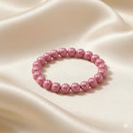 Ein Armbands aus glänzenden, rosa Perlen, das auf einem beigefarbenen, seidenartigen Stoff liegt.