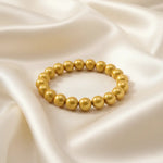 Ein Armbands aus glänzenden, goldfarbenen Perlen, das auf einem beigefarbenen, seidenartigen Stoff liegt.