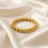 Ein Armbands aus glänzenden, goldfarbenen Perlen, das auf einem beigefarbenen, seidenartigen Stoff liegt.