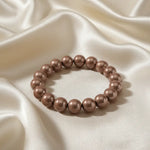 Ein Armbands aus glänzenden, taupefarbenen Perlen, das auf einem beigefarbenen, seidenartigen Stoff liegt.