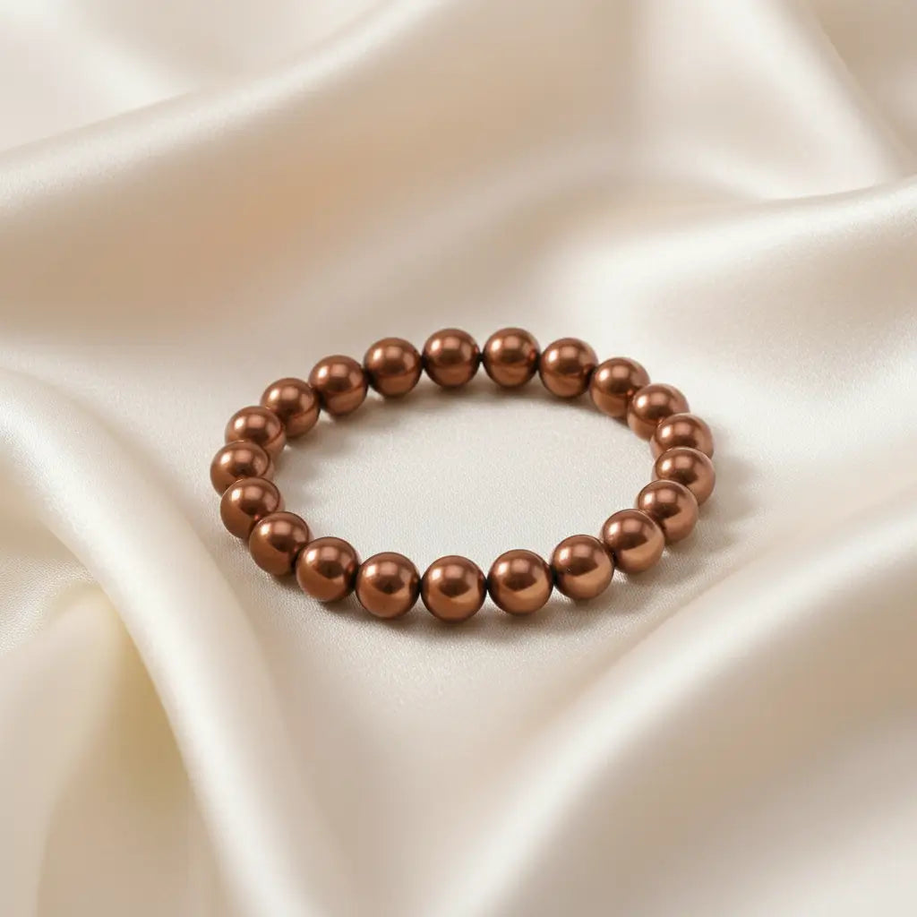 Ein Armbands aus glänzenden, braunen Perlen, das auf einem beigefarbenen, seidenartigen Stoff liegt.