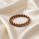 Ein Armbands aus glänzenden, braunen Perlen, das auf einem beigefarbenen, seidenartigen Stoff liegt.