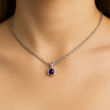 Nahaufnahme eines Frauenhalses mit einer silbernen Kette und einem kleinen, runden Amethyst-Cabochon-Anhänger von 5 mm Durchmesser. Der Anhänger zeigt einen tiefvioletten, glatten Stein in einer polierten Silberfassung. Der Hintergrund ist neutral und weich ausgeleuchtet, der Fokus liegt klar auf dem Anhänger.