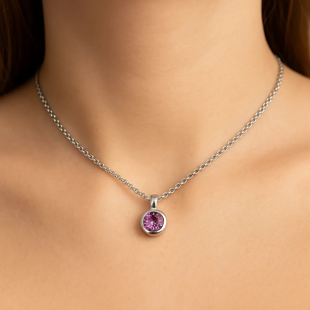 Nahaufnahme eines Frauenhalses mit einer silbernen Kette und einem kleinen, runden, facettierten Amethyst-Anhänger von 5 mm Durchmesser. Der violett funkelnde Edelstein ist in einer glatten Silberfassung eingefasst. Der Hintergrund ist neutral und weich ausgeleuchtet, der Fokus liegt auf dem Anhänger.