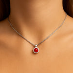 Nahaufnahme eines Frauenhalses mit einer silbernen Kette und einem kleinen, runden Korallen-Cabochon-Anhänger von 5 mm Durchmesser. Der leuchtend rote, glatte Stein ist in einer glänzenden Silberfassung eingefasst. Der Hintergrund ist neutral und weich ausgeleuchtet, der Fokus liegt auf dem Anhänger.