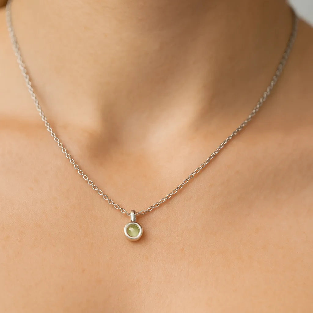 Nahaufnahme eines Frauenhalses mit einer feinen silbernen Kette und einem kleinen, runden Peridot-Cabochon-Anhänger von 5 mm Durchmesser. Der grün schimmernde Stein ist in Silber gefasst, der Hintergrund ist neutral und weich ausgeleuchtet, der Fokus liegt auf dem dezenten Anhänger.