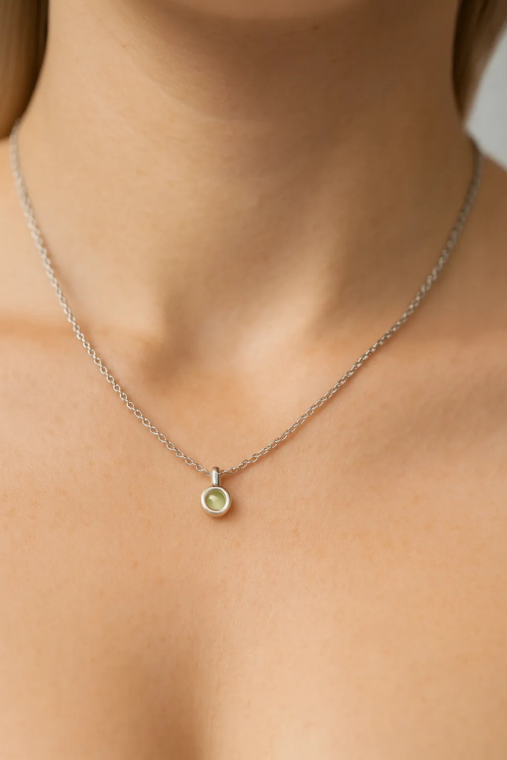 Nahaufnahme eines Frauenhalses mit einer feinen silbernen Kette und einem kleinen, runden Peridot-Cabochon-Anhänger von 5 mm Durchmesser. Der grün schimmernde Stein ist in Silber gefasst, der Hintergrund ist neutral und weich ausgeleuchtet, der Fokus liegt auf dem dezenten Anhänger.