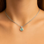 Nahaufnahme eines Frauenhalses mit einer silbernen Kette und einem kleinen, runden Prehnit-Cabochon-Anhänger von 5 mm Durchmesser. Der zartgrüne, leicht durchscheinende Stein ist in einer glatten Silberfassung eingefasst. Der Hintergrund ist neutral und weich ausgeleuchtet, der Fokus liegt auf dem Anhänger.