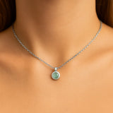 Nahaufnahme eines Frauenhalses mit einer silbernen Kette und einem kleinen, runden Prehnit-Cabochon-Anhänger von 5 mm Durchmesser. Der zartgrüne, leicht durchscheinende Stein ist in einer glatten Silberfassung eingefasst. Der Hintergrund ist neutral und weich ausgeleuchtet, der Fokus liegt auf dem Anhänger.