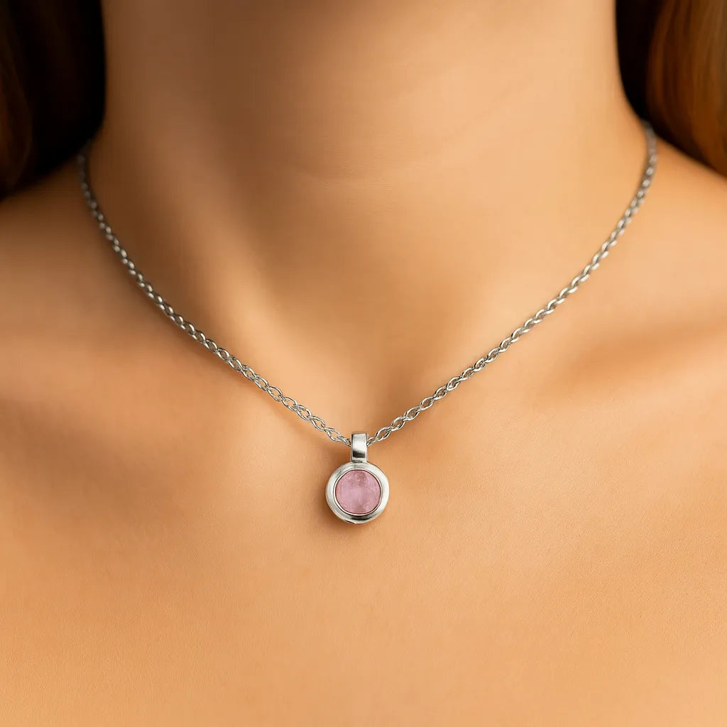 Nahaufnahme eines Frauenhalses mit einer silbernen Kette und einem kleinen, runden Rosenquarz-Anhänger von 5 mm Durchmesser. Der zartrosa, leicht trübe facettierte Stein ist in einer glänzenden Silberfassung eingefasst. Der Hintergrund ist neutral und weich ausgeleuchtet, der Fokus liegt auf dem Anhänge
