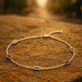 Ein silbernes Armband mit fünf rechteckigen, farbigen Edelsteinen liegt auf einem sonnenbeschienenen Stein in einer urbanen Umgebung. Im Hintergrund sind unscharfe Gebäude und warmes, goldenes Abendlicht zu sehen, das die funkelnden Steine stimmungsvoll betont.