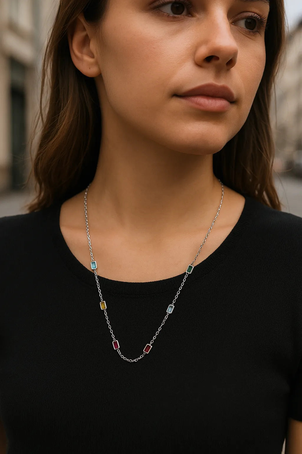 Eine junge Frau steht in einer urbanen Umgebung und trägt eine zarte Silberkette mit rechteckigen Edelsteinen in den Farben Hellblau, Grün, Gelb, Hellblau und zwei Rottönen. Die Kette liegt elegant auf einem schwarzen Oberteil und wird durch das natürliche Licht dezent hervorgehoben. Der Hintergrund ist leicht unscharf, wodurch der Fokus klar auf der Halskette liegt.