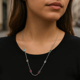 Eine junge Frau steht in einer urbanen Umgebung und trägt eine zarte Silberkette mit rechteckigen Edelsteinen in den Farben Hellblau, Grün, Gelb, Hellblau und zwei Rottönen. Die Kette liegt elegant auf einem schwarzen Oberteil und wird durch das natürliche Licht dezent hervorgehoben. Der Hintergrund ist leicht unscharf, wodurch der Fokus klar auf der Halskette liegt.
