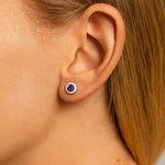 Nahaufnahme einer Frau im Profil mit einem kleinen runden Ohrstecker aus Silber und einem violetten Amethyst im Ohr. Der facettierte Edelstein funkelt im Licht, während die glatte, helle Haut und das zurückgelegte blonde Haar den Ohrring deutlich hervorheben