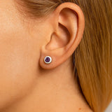 Nahaufnahme einer Frau im Profil mit einem kleinen runden Ohrstecker aus Silber und einem violetten Amethyst im Ohr. Der facettierte Edelstein funkelt im Licht, während die glatte, helle Haut und das zurückgelegte blonde Haar den Ohrring deutlich hervorheben