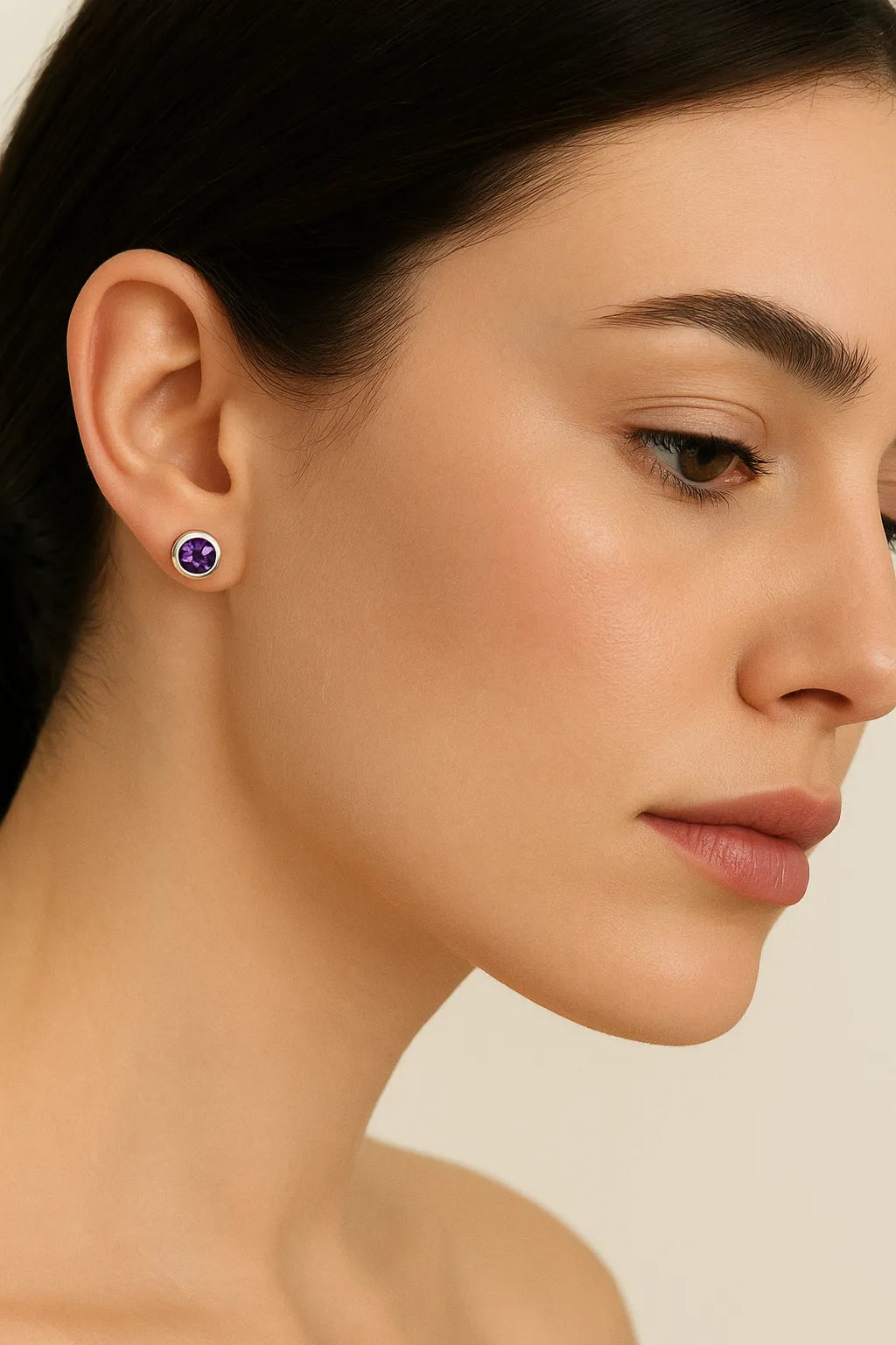 Porträt einer Frau in Seitenansicht, die einen silbernen Ohrstecker mit einem runden, facettierten Amethyst trägt. Der violette Edelstein funkelt dezent im Licht, während die Frau mit glatter Haut und dezentem Make-up vor einem neutralen Hintergrund zu sehen ist.