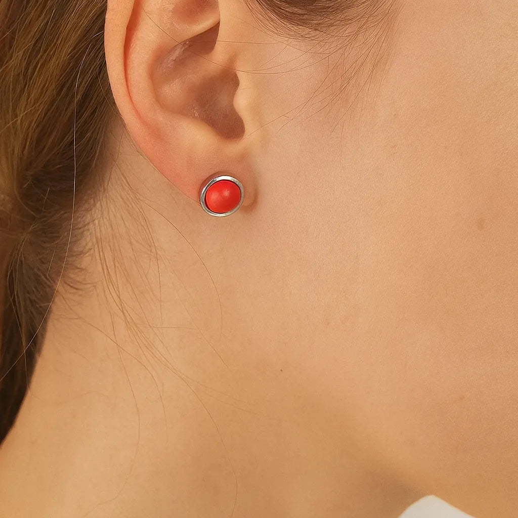 Nahaufnahme einer Frau in Seitenansicht mit einem kleinen silbernen Ohrstecker, der eine glänzende, korallenrote Kuppel zeigt. Der Ohrring hat einen Durchmesser von etwa 5 mm und sitzt dezent am Ohrläppchen, während Haar und Haut in warmem Licht natürlich wirken.