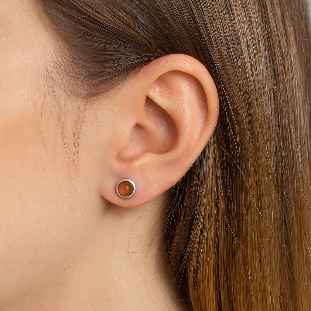 Nahaufnahme eines weiblichen Ohrs mit einem eleganten Ohrstecker aus 925er Sterlingsilber, besetzt mit einem runden orangefarbenen Mondstein-Cabochon. Der warme Glanz des Steins verleiht dem Schmuckstück eine natürliche, dezente Eleganz.