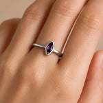 Nahaufnahme einer Frauenhand, die einen eleganten Silberring mit einem kleinen, länglich geschliffenen Amethyst trägt. Der violette Edelstein sitzt mittig in einer zarten Ringschiene und ist etwa 5 mm lang, wodurch er dezent und edel wirkt. Der Fokus des Bildes liegt klar auf dem Ring, während der Hintergrund neutral und weich gehalten ist.
