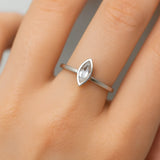 Eine Nahaufnahme einer Frauenhand, die einen eleganten Silberring mit einem kleinen, länglich facettierten Mondstein am Ringfinger trägt. Der transparente Stein zeigt zarte Blauschimmer, eingefasst in eine schlichte, glänzende Fassung. Der Fokus liegt klar auf dem Ring, während der Hintergrund neutral und weich verschwommen ist.
