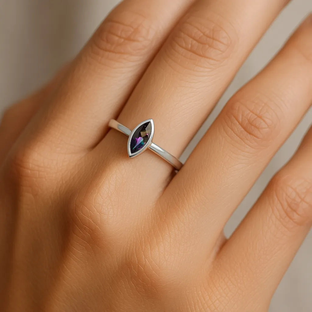 Eine Nahaufnahme einer weiblichen Hand, die einen eleganten Silberring mit einem marquisförmigen Mystic-Topas trägt. Der Stein zeigt ein schimmerndes Farbspiel in Violett-, Grün- und Blautönen und misst etwa 5 mm. Der Fokus liegt auf dem Ring, während die Haut weich ausgeleuchtet und der Hintergrund neutral gehalten ist.

