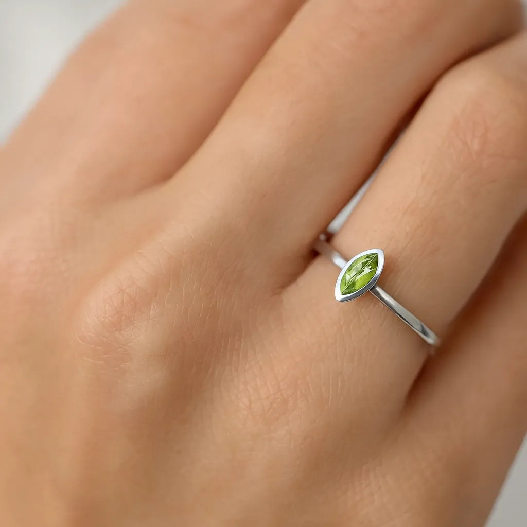 Nahaufnahme einer Frau, die einen eleganten Silberring mit einem 5 mm langen, marquiseförmig geschliffenen Peridotstein am Ringfinger trägt. Der Ring hat ein schlichtes, glänzendes Design mit dem grünen Edelstein als zentralem Blickfang. Der Fokus liegt auf dem Ring, während die Haut der Hand weich ausgeleuchtet ist.